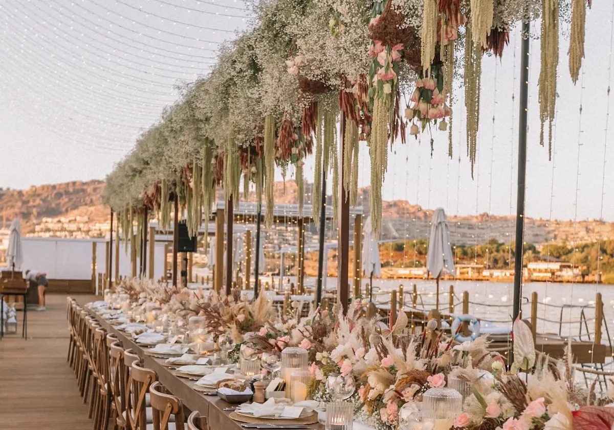 Bodrum'un Ege kıyısında gün batımına doğru uzanan bir iskele üzerinde, zarif ahşap masaların pastel tonlarda pembe güller, pampas otları ve sarkan yeşil bitkilerle donatıldığı, üzerinde ışıklandırmalar ve cipsolarla süslenmiş tavanıyla kusursuz bir düğün organizasyonu için hazırlanmış açık hava kutlaması alanı.