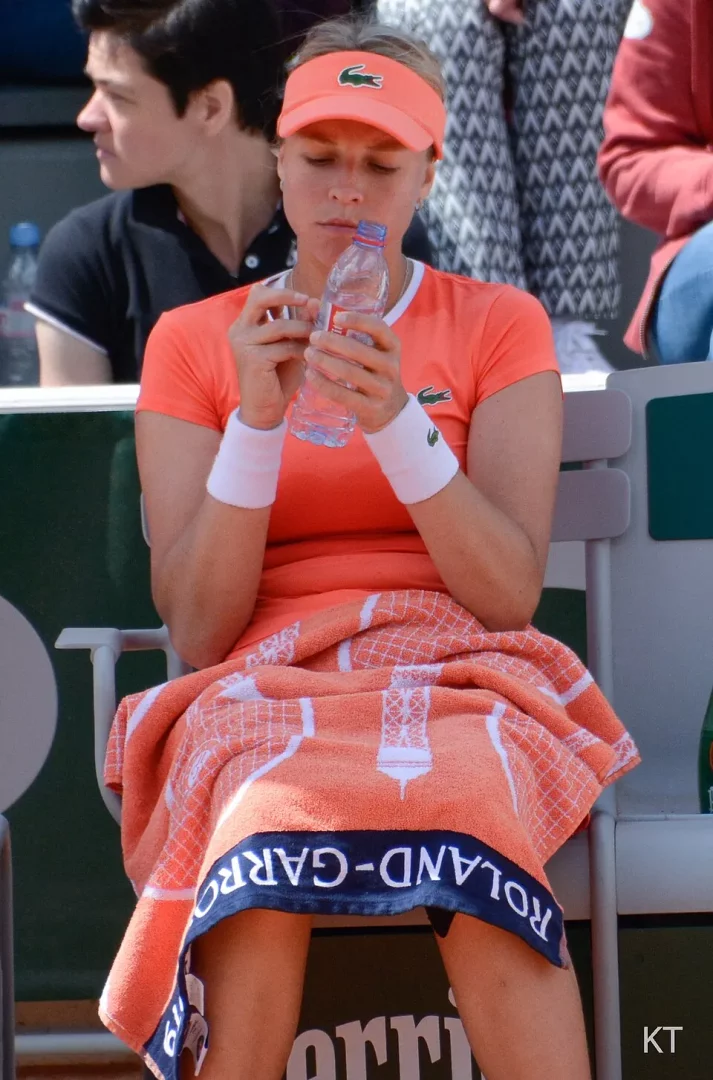 Orange siperlikli şapkası, Lacoste logolu tişörtü ve beyaz bileklikleriyle, kucağında Roland Garros logolu turuncu bir havlu bulunan tenisçi Karolina Muchova, kort kenarında dinlenirken elindeki su şişesine dikkatle bakarak susuzluğunu gidermeye hazırlanıyor.