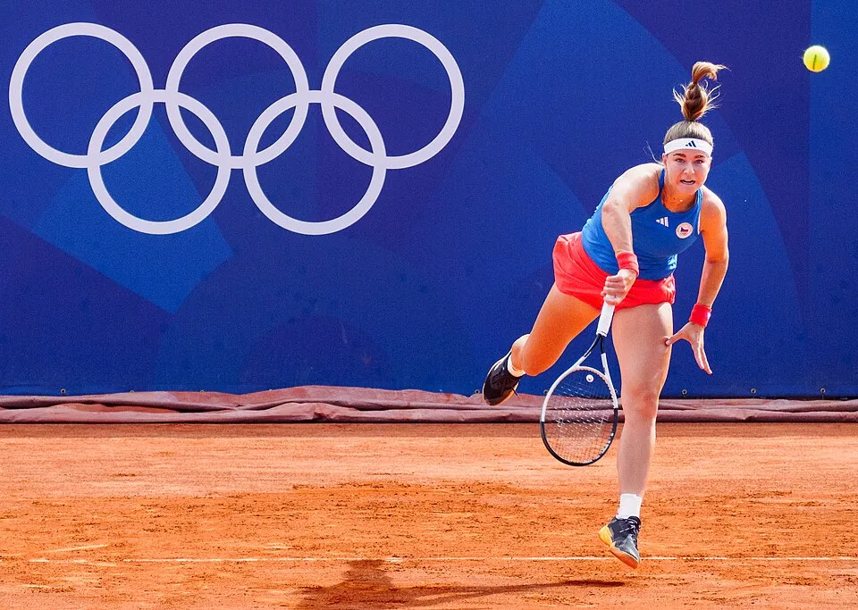 Karolina Muchova, 2024 Olimpiyatları'na hazırlanırken, Çek Cumhuriyeti bayraklı mavi tişörtü ve kırmızı şortuyla toprak kortta, havada süzülen tenis topuna enerjik bir smaç vuruşu yapıyor; arka planda Olimpiyat halkaları belirgin şekilde seçiliyor.
