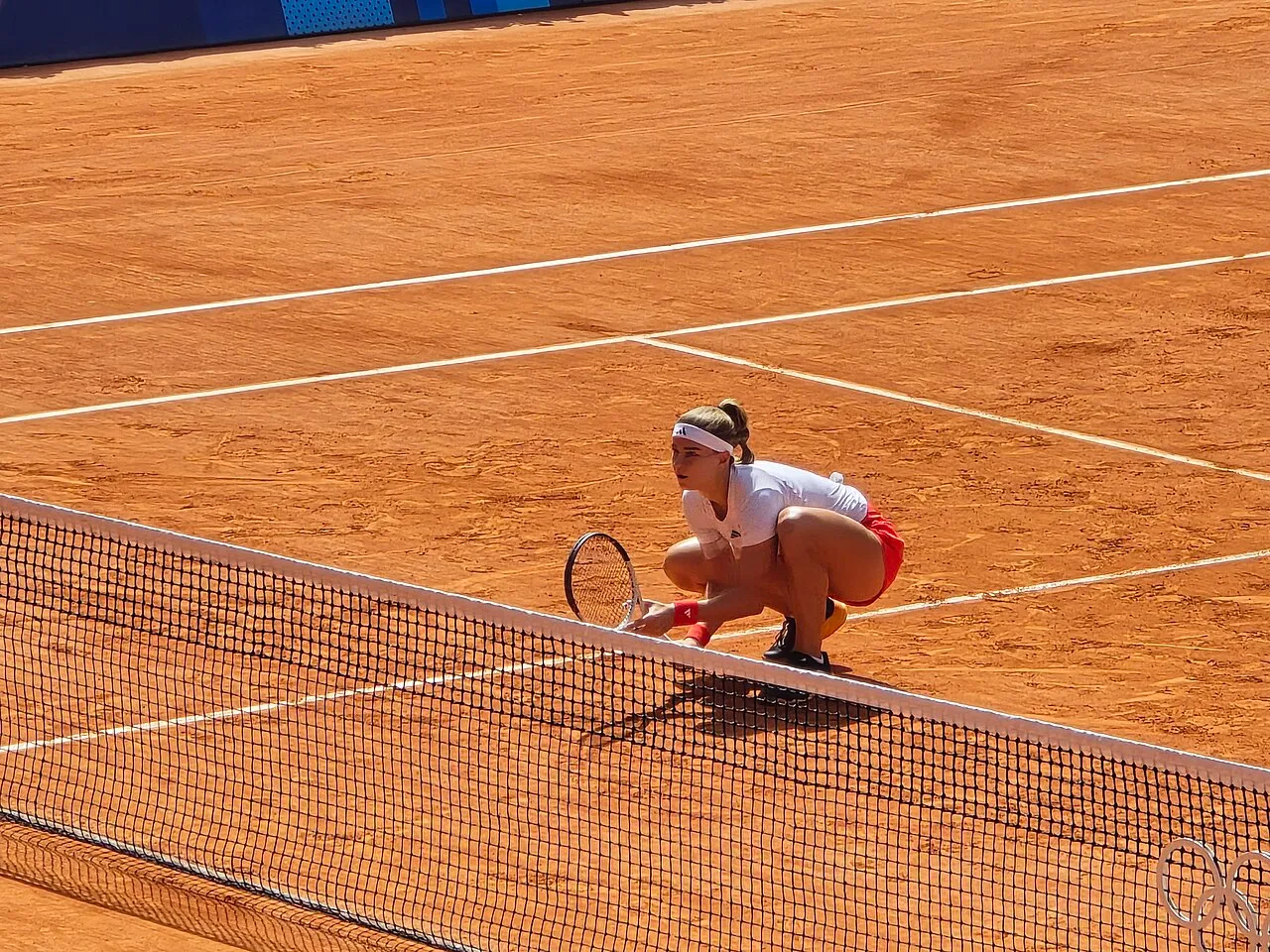 2024 Olimpiyatları tenis turnuvası kapsamında kızıl toprak kortta, fileye yakın bir noktada beyaz tişört ve kırmızı şortuyla çömelmiş, raketini yere dayamış Karolina Muchova, dikkatle etrafına bakarken izleniyor.