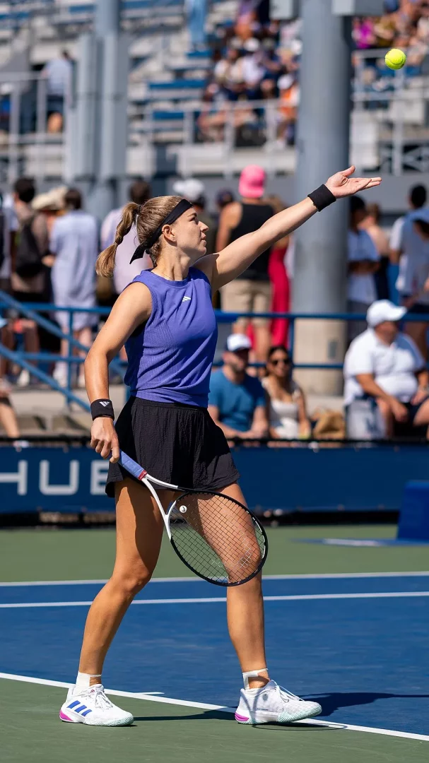 2024 US Open tenis turnuvasında Karolina Muchova, mavi ve yeşil zeminli bir kortta mor atlet ve siyah etekle servis atmaya hazırlanırken, tenis topunu sol eliyle havaya fırlatmış, raketi sağ elinde tutarken arka plandaki tribünlerde kalabalık seyirciler görülüyor.