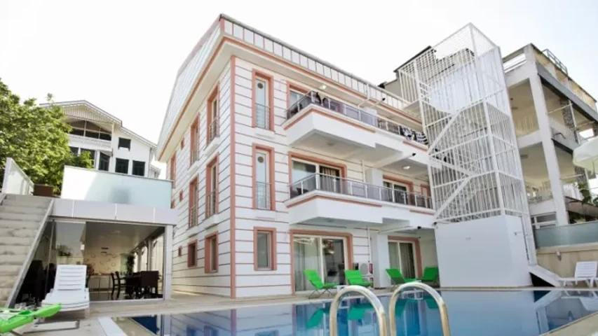 Beyaz ve açık şeftali renklerinde dış cephesi olan, geniş cam balkonlu, modern bir Kınalıada otelinin önünde, berrak mavi suya sahip bir yüzme havuzu ve etrafına dizilmiş yeşil şezlonglar görünüyor, bu huzurlu tesisin ferahlatıcı yaz atmosferini vurguluyor.