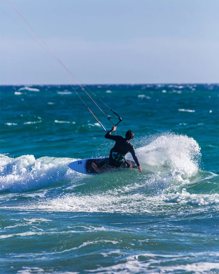 Mavi gökyüzü altında berrak turkuaz denizde, siyah wetsuit giyen bir kitesurf sporcusu, enerjik bir dalga üzerinde sörf tahtasıyla ilerleyerek çevresine beyaz köpüklü sular sıçratırken heyecan verici bir uçurtma sörfü deneyimi yaşıyor.