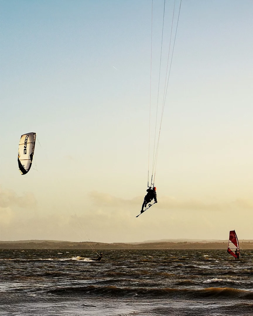 Gün batımının altın rengi ışıkları altında, dalgalı denizde bir kitesurfçü tüm ekipmanlarıyla havada süzülerek adeta denizin üzerinden yükseliyor; sağda kırmızı yelkenli bir rüzgar sörfçüsü, sol arkada ise bir başka kitesurfçü su üzerinde ilerliyor.