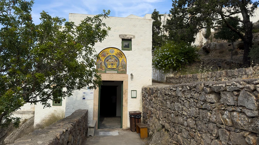 Kıyamet Adası Patmos Adası'nda, kemerli giriş kapısının üzerinde altın rengi dini figürlerle bezenmiş görkemli bir mozaiğin yer aldığı beyaz badanalı tarihi bir yapı, sağında yükselen taş duvarlı patika ve yemyeşil ağaçlarla çevrili bir ortamda görülüyor.