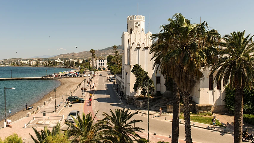 Güneşli bir Kos Adası gününde, sağda karakteristik beyaz saat kuleli belediye binasının yükseldiği, palm ağaçlarıyla süslü, hareketli sahil yolu, solunda çakıllı plajı ve masmavi deniziyle uzanır; uzakta dağlar belirir.