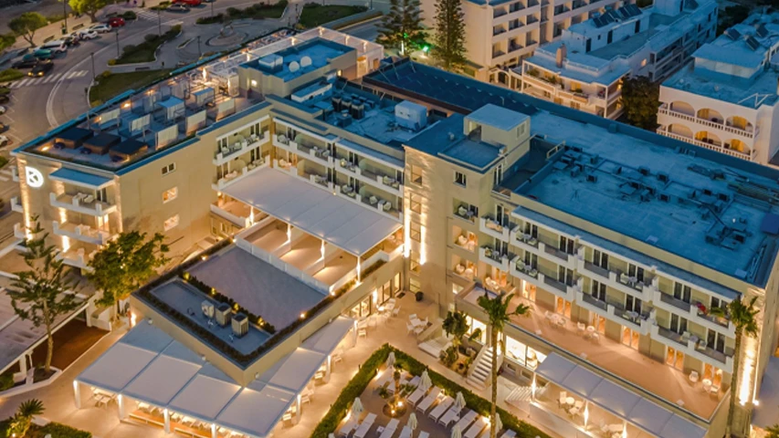 Alacakaranlıkta çekilmiş bu genel bakış fotoğrafında, Kos Divine Hotel Suites'in şık mimarisi, aydınlatılmış yüzme havuzu, düzenli şezlongları ve yemyeşil palmiyelerle çevrili açık hava alanları, otelin lüks ve davetkar atmosferini yansıtırken çevresindeki yol ve binalar da görülmektedir.