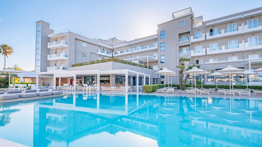 Gün ışığında parlayan modern Kos Divine Hotel Suites binası, önünde uzanan berrak mavi yüzme havuzu ve çevresindeki şemsiyeli şezlonglarla huzurlu bir atmosfer sunarken, palmiye ağaçları ve açık hava bar alanı dinlenmeye davet ediyor.