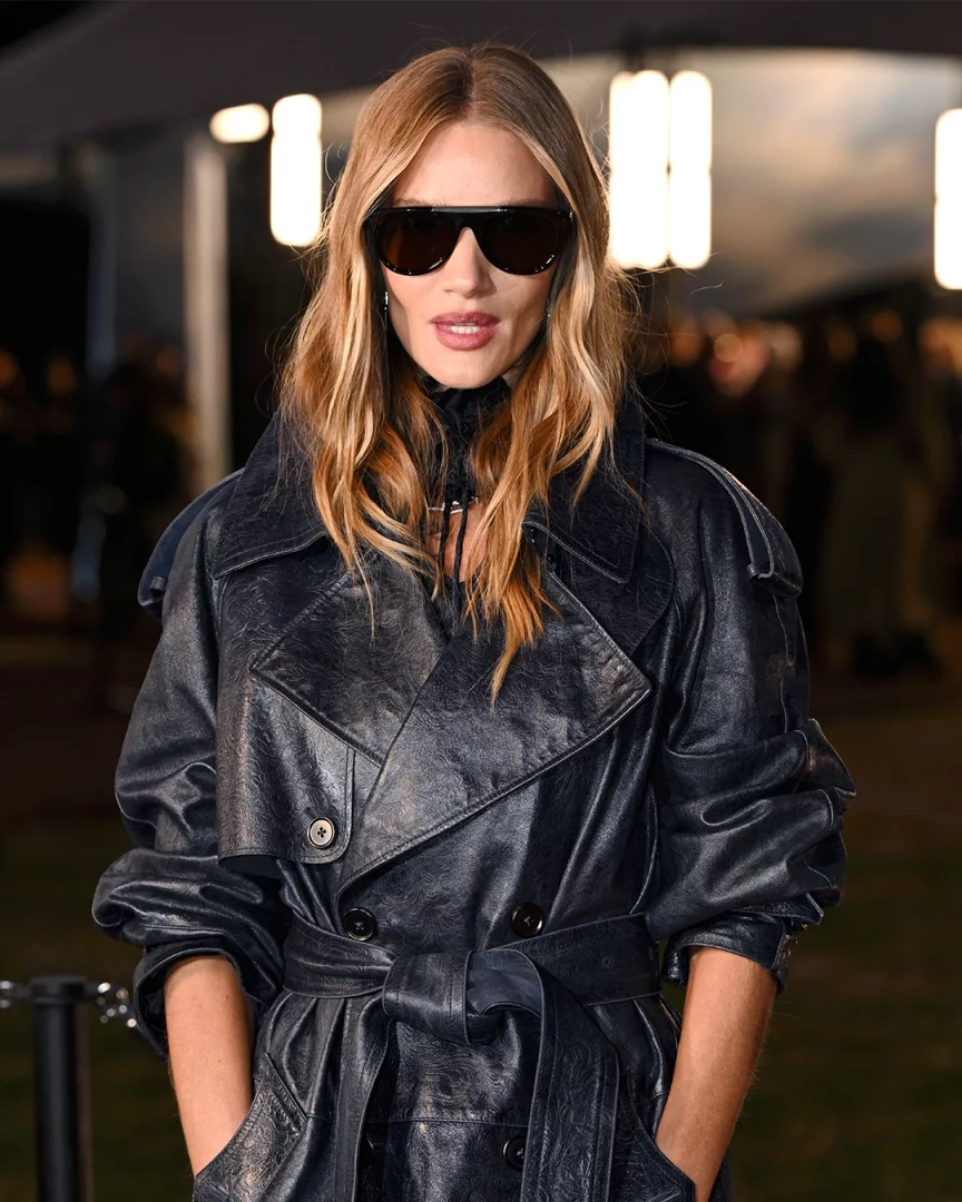 Rosie Huntington-Whiteley, dalgalı ve kumral-sarı (bronde) saçları, büyük siyah güneş gözlükleri ve kemerli parlak siyah deri trençkotuyla elleri ceplerinde objektife bakarken şık bir poz veriyor, arkasında bulanık gece ışıkları görünüyor.