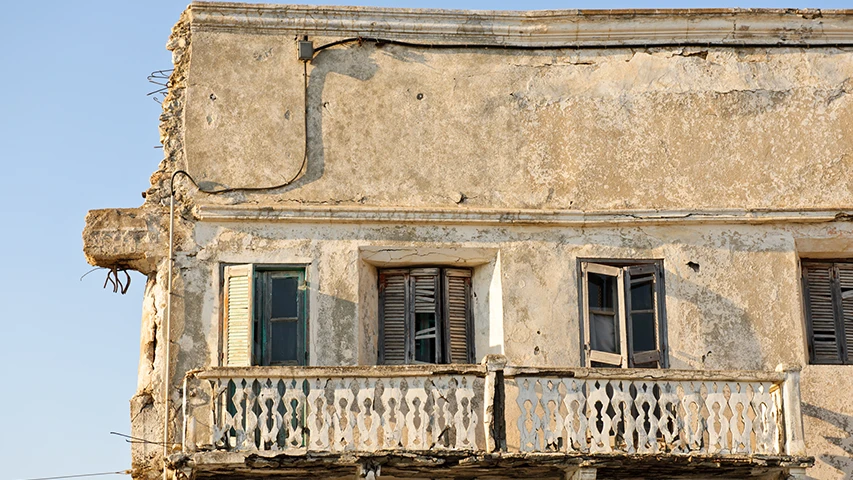 Leros'un Lakki kasabasındaki bir yapının yakından çekilmiş bu fotoğrafında, sıvaları dökülmüş, betonarme demirleri görünen yıpranmış bir duvar, farklı renk ve açıklıktaki eski ahşap kepenkli pencereler ve işlemeli, aşınmış bir balkon korkuluğu dikkat çekerek bölgenin kendine özgü yaşanmışlığını ve dokusunu vurguluyor.