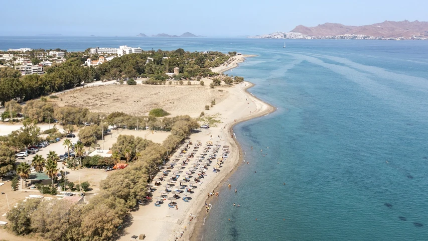 Güneşli bir günde Kos Adası'ndaki Lambi Plajı'nın kuşbakışı görüntüsü, berrak turkuaz sularında yüzen ve geniş kumlu plajında şemsiyelerin altında dinlenen birçok insanı gösteriyor; kıyı boyunca oteller ve yeşil alanlar uzanırken, ufukta uzanan dağlık arazi manzarayı tamamlıyor.