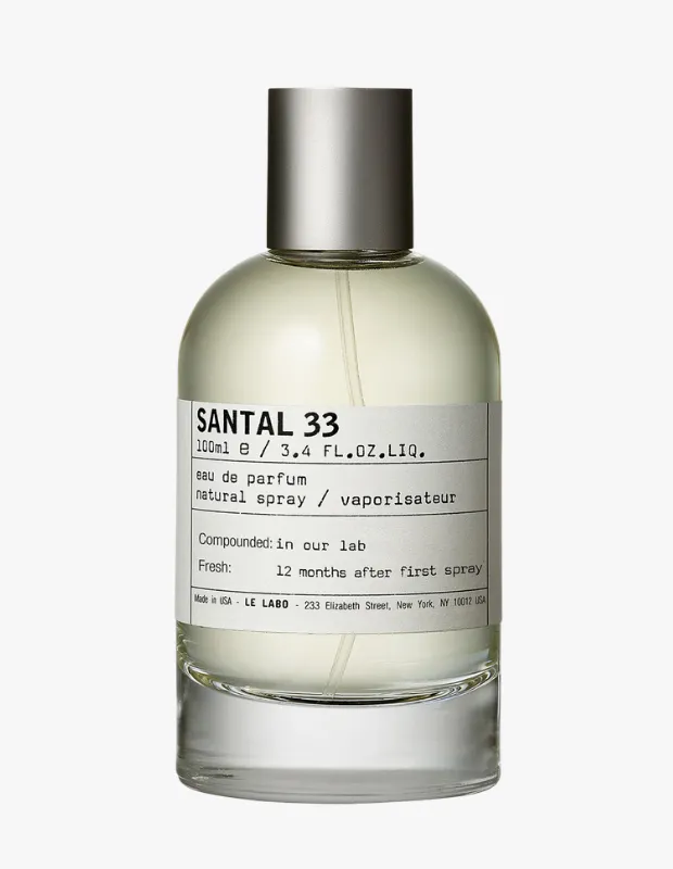 Parlak beyaz bir zeminde dikey duran, Le Labo Santal 33 etiketli, 100 ml'lik şeffaf cam şişedeki eau de parfum, gümüş renkli metalik bir kapak ve ürünün içeriği ile doğal sprey bilgilerini içeren beyaz bir etikete sahiptir.