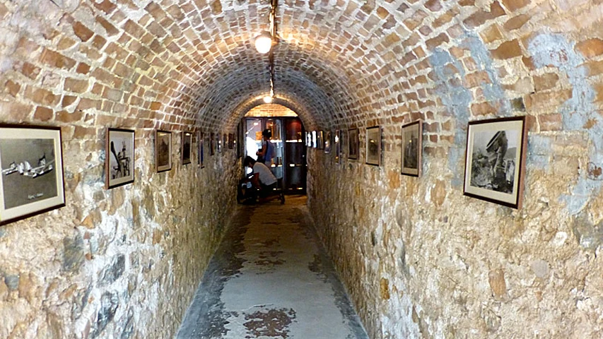 Leros Savaş Müzesi'nde, eski taş ve tuğla duvarların oluşturduğu kemerli bir tünelin iki yanında, geçmişe dair karelerin yer aldığı sıralı çerçeveli siyah beyaz fotoğraflar loş ışıklar altında sergilenirken, ileride birkaç ziyaretçi dikkatle etrafa bakıyor.