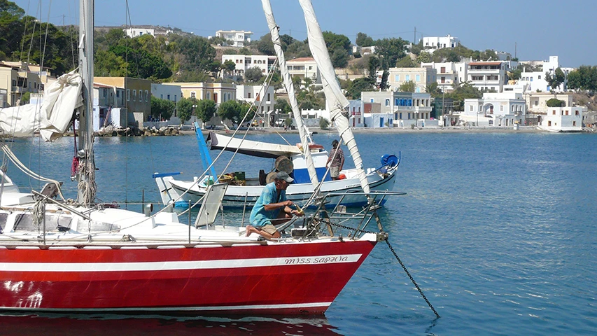 Leros limanında demirli, üzerinde çalışan bir adamın olduğu kırmızı yelkenli tekne ve arkadaki şirin sahil kasabası manzarası, adanın taze deniz ürünleri ve yöresel lezzetlerinin tadına bakmak için ne yenir sorusuna ilham veriyor.