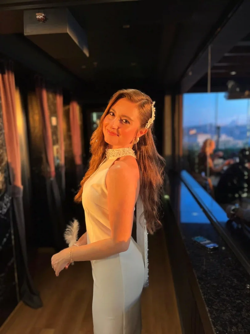 Lidya Amun, mutlu bir hikâyenin başlangıcı olan bekarlığa veda kutlamasında inci temalı beyaz, yüksek yakalı elbisesi ve incili saç aksesuarıyla Pera Adalı Restaurant'ın panoramik şehir manzaralı loş ışıklı ortamında neşeyle gülümsüyor.