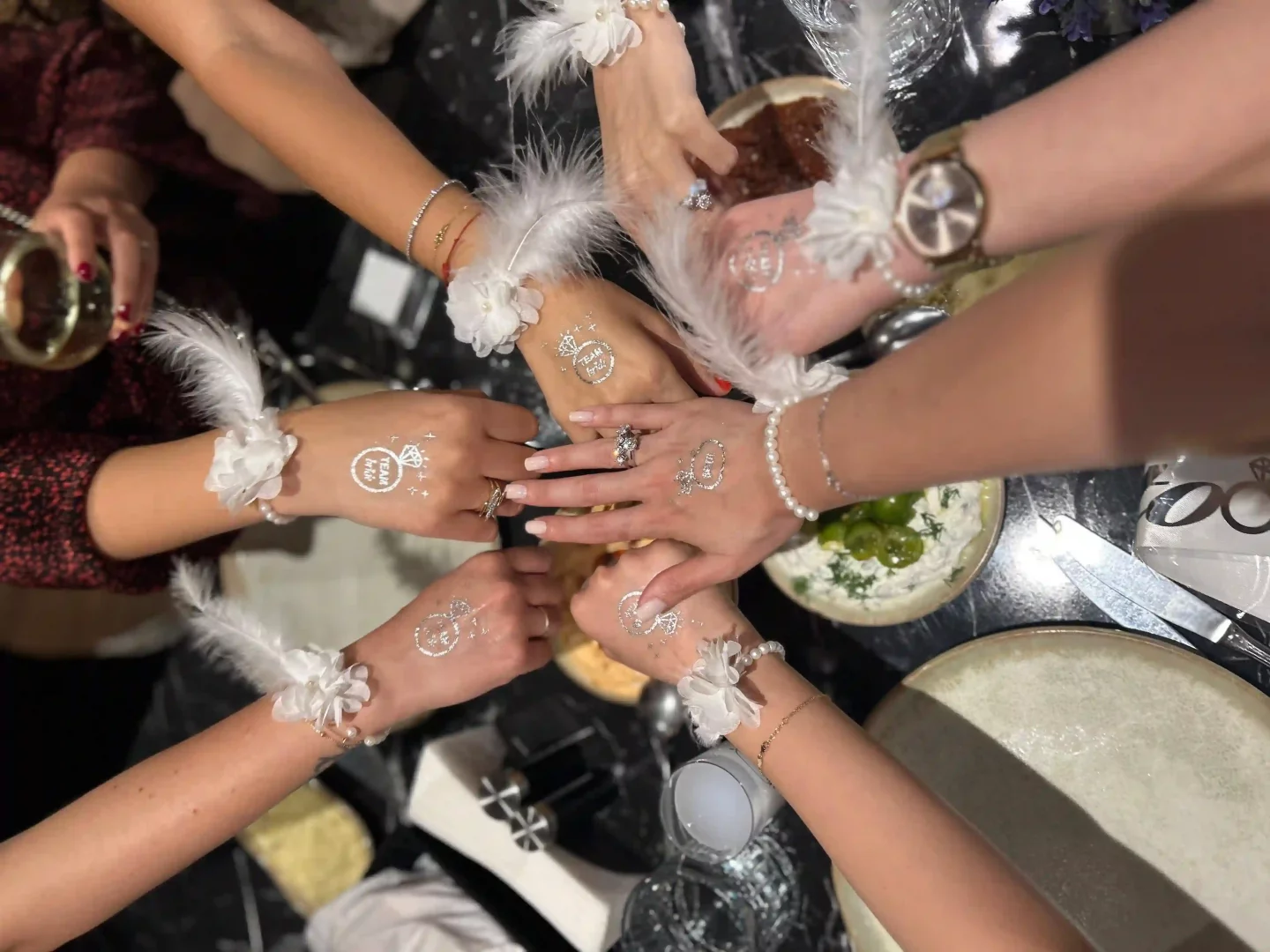 Bir daire şeklinde bir araya gelmiş kadın elleri, üzerinde beyaz tüy detaylı bileklikler ve bazı ellerde pırlanta yüzük figürlü TEAM BRIDE yazan gümüş renkli geçici dövmelerle neşeli bir bekârlığa veda kutlaması anını yansıtarak mutlu bir hikâyenin başlangıcına vurgu yapıyor.