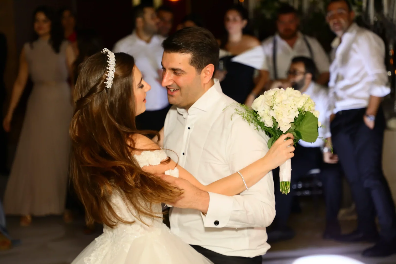 Lidya Amun ve Cem Amun'un düğünlerindeki ilk dansları sırasında çekilen bu romantik karede, kabarık gelinliği ve kristal tacıyla Lidya elinde beyaz çiçeklerden oluşan gelin buketiyle eşine sevgiyle bakarken, Cem Amun da gömleğiyle eşini nazikçe kucaklayarak neşeyle gülümsüyor.