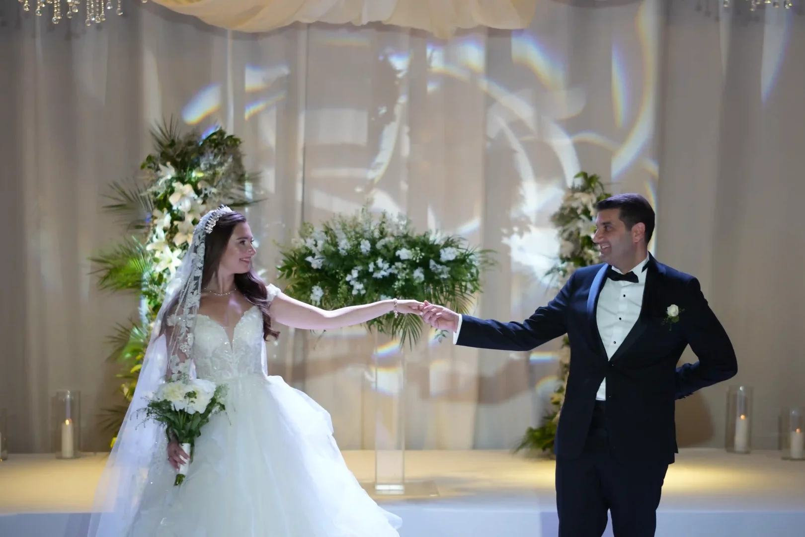 Lidya ve Cem Amun, beyaz şakayık buketi tutan Lidya'nın kabarık gelinliği ve Cem'in klasik smokiniyle, beyaz çiçek ve yeşilliklerle süslü zarif bir düğün salonunda el ele ilk danslarını sergilerken, iki geleneği buluşturan aşklarının başlangıcını neşeyle kutluyorlar.
