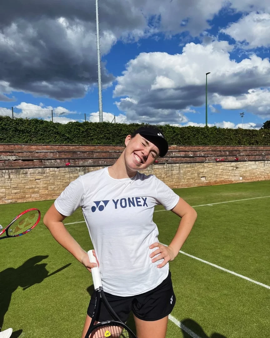 Linda Nosková, Yonex logolu beyaz tişörtü ve siyah şortuyla, elinde tenis raketiyle güneşli bir çim tenis kortunda gülümseyerek poz veriyor; arka planda tuğla tribünler, yeşil çalılar ve bulutlu mavi gökyüzü dikkat çekiyor.