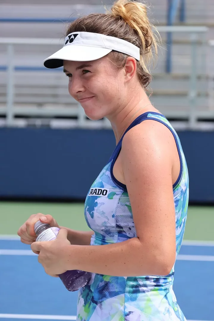 Genç tenis oyuncusu Linda Noskova, dağınık topuz yapılmış saçları, mavi-yeşil-beyaz desenli RADO logolu tenis kıyafeti ve beyaz YONEX vizörüyle tenis kortunda, elindeki mor renkli içecek şişesini açmaya çalışırken sağ profilden hafifçe gülümsüyor.