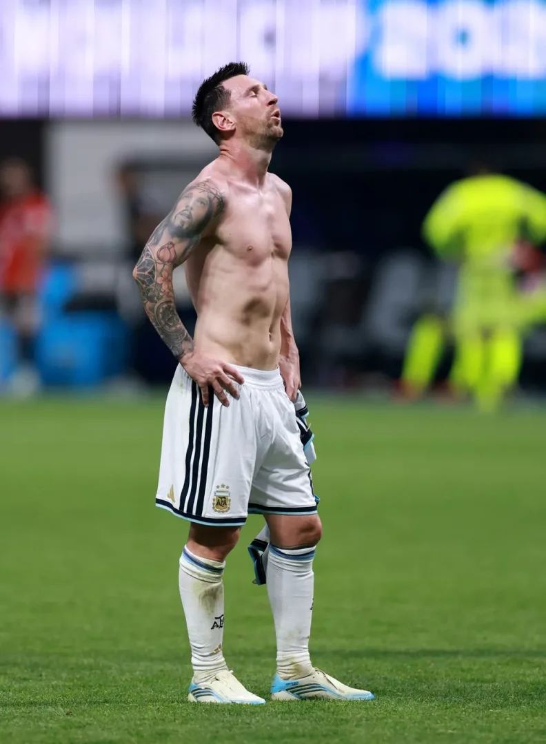 Çim sahada, forma üstünü çıkarmış, dövmeli sol kolu ve belirgin kaslarıyla Lionel Messi, elleri belinde, yorgun ve düşünceli bir ifadeyle başını yukarı kaldırmış, 2026 Dünya Kupası ihtimali ve kariyerinin uzun ömürlülüğü üzerine kafa yoruyor gibi görünüyor.