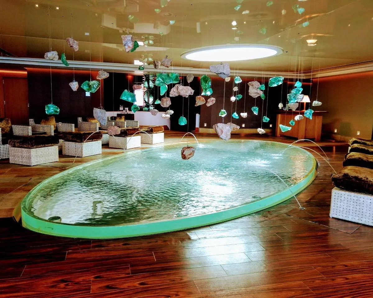 Uzun ömürlülük temalı bir spa merkezinde, şeffaf ve doğal görünümlü taşların asılı olduğu yüksek tavanlı, pırıl pırıl ahşap zeminli geniş bir alanda, su fışkırtan oval havuz ve çevresindeki beyaz dokulu, konforlu oturma grupları dinlendirici bir ortam sunuyor.