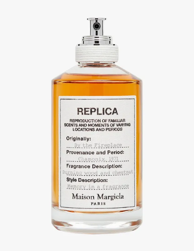 Beyaz bir zeminde sergilenen, kehribar rengi sıvısı ve gümüş püskürtücüsüyle Maison Margiela Replica By the Fireplace parfüm şişesi, yanan odun ve kestane kokusu ile bir anıdaki koku tanımını içeren ikonik beyaz etiketiyle detaylıca görünüyor.