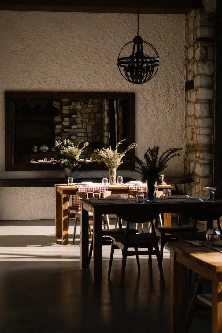 Urla'daki Manej restoranının güneş ışığı alan iç mekanında, rustik ahşap masalar, ekose örtüler, kurutulmuş bitkilerle süslü vazolar, aynadaki yansımalar ve taş duvar detaylarıyla sıcak bir atmosfer sunuluyor.