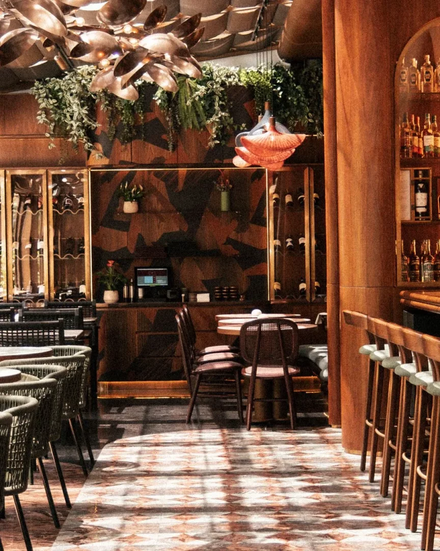 Modern ve davetkar bir atmosfer sunan bir restoran veya barın iç mekanında, şık ahşap panellerin üzerine asılmış yemyeşil bitkiler ve çarpıcı bronz avizelerle zenginleştirilmiş tavan, farklı oturma düzenlerine sahip masalar ve yüksek bar sandalyeleriyle dolu, karmaşık desenli mermer zeminde ışık yansımalarının dans ettiği sıcak ve detaylı bir tasarım.