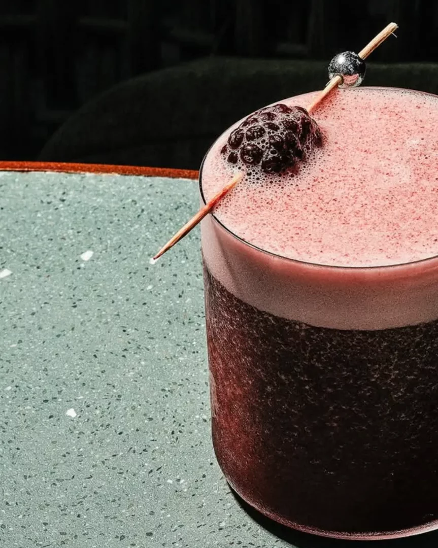 İstanbul'daki Manze restoranında sunulan, koyu kırmızı renkte, üzerinde pembe köpük tabakası bulunan ve ahşap çubuğa takılı parlak bir böğürtlen ile gümüş rengi bir boncukla zarifçe süslenmiş serinletici bir kokteylin yakından çekilmiş görüntüsü, yeşil benekli bir masanın üzerinde duruyor.