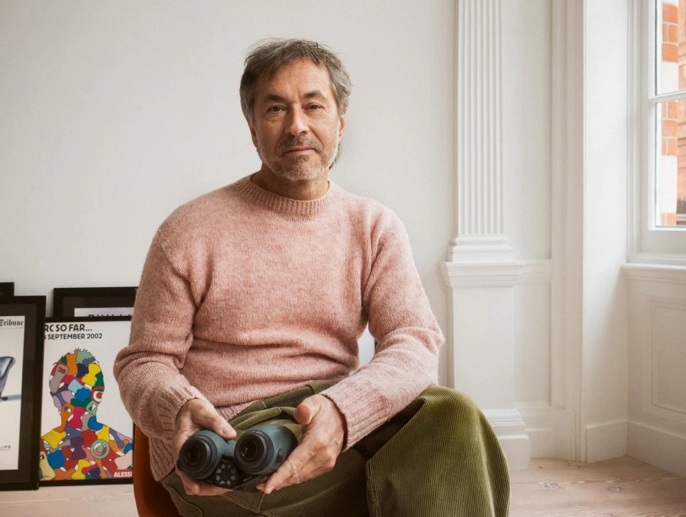 Tasarımlarıyla tanınan Marc Newson, açık pembe örgü kazağı ve zeytin yeşili kadife pantolonuyla sade bir odada oturmuş, ellerinde yeşil renkli bir dürbün tutarak doğrudan objektife bakıyor; arkasında çerçeveli sanat eserleri ve dışarıyı gösteren bir pencere seçiliyor.