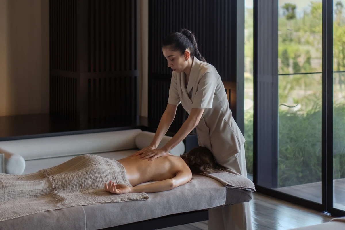 Lüks bir spa ortamında, masaj yatağında yüzüstü uzanmış bir müşteriye sırt masajı yapan üniformalı bir terapist görülüyor; pencereden dışarıdaki yeşil bitki örtüsü huzurlu bir atmosfer sunarak, dinlendirici bir spa masaj deneyimi yaşatıyor.
