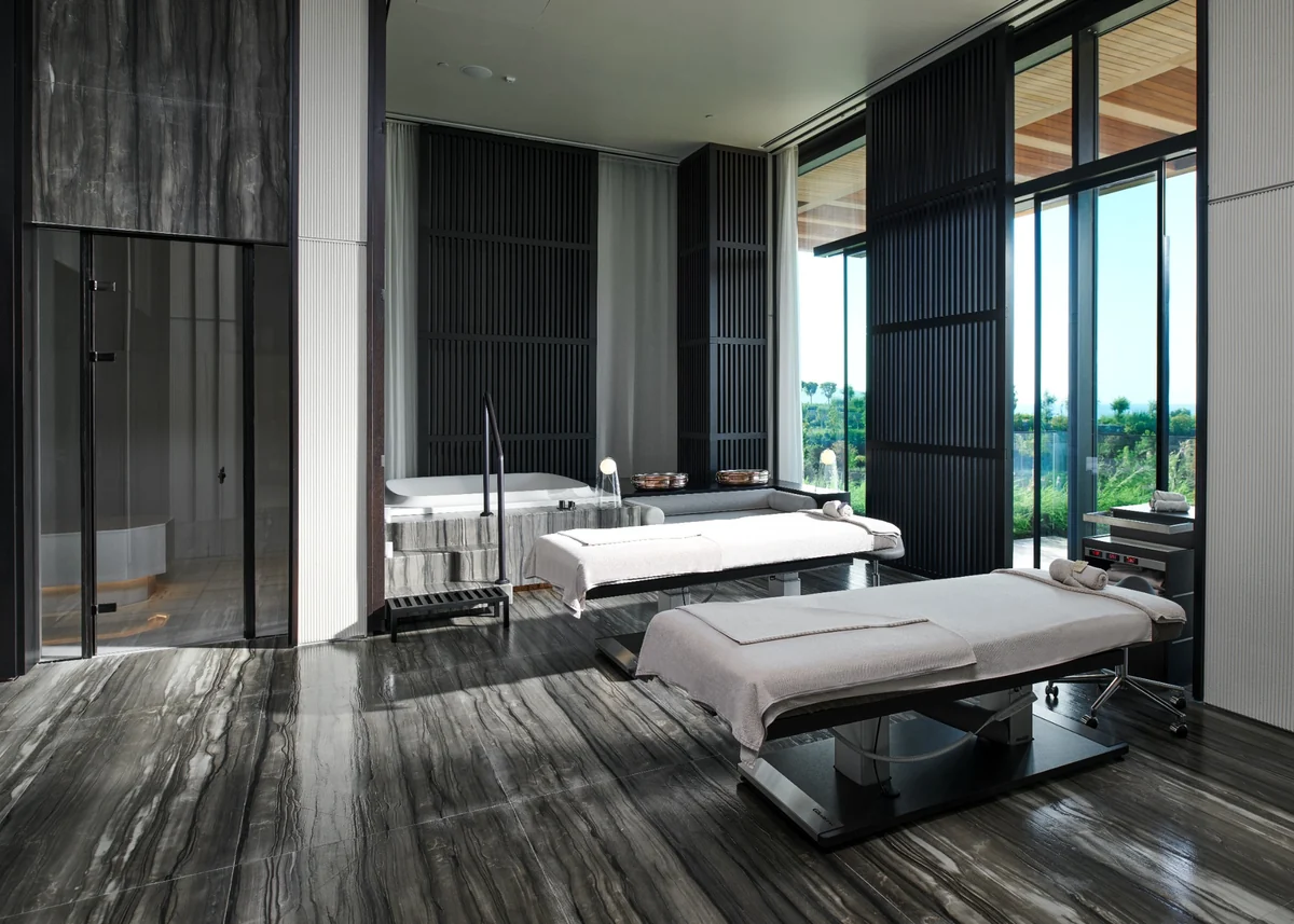 Maxx Royal Bodrum'da lüks otelciliğin wellness trendi kapsamındaki bütünsel yenilenme deneyimlerini temsil eden, koyu mermer zeminli, iki beyaz örtülü masaj masası ve yüksek platformda bir küvetin bulunduğu, geniş camlarından yeşil doğanın göründüğü, modern ve dingin bir spa terapi odası.