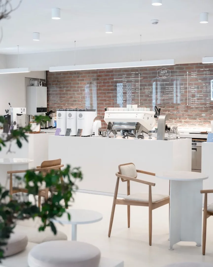 Meet Lab Coffee'nin aydınlık ve modern iç mekanında, parlak beyaz bir servis tezgahı üzerinde ileri teknoloji kahve ekipmanları dikkat çekerken, arkadaki tuğla duvar önünde markanın menüsünü sergileyen şeffaf bir panel ve ön planda açık renk ahşap sandalyeler ile minimalist küçük beyaz masalar ferah bir oturma alanı sunmaktadır.