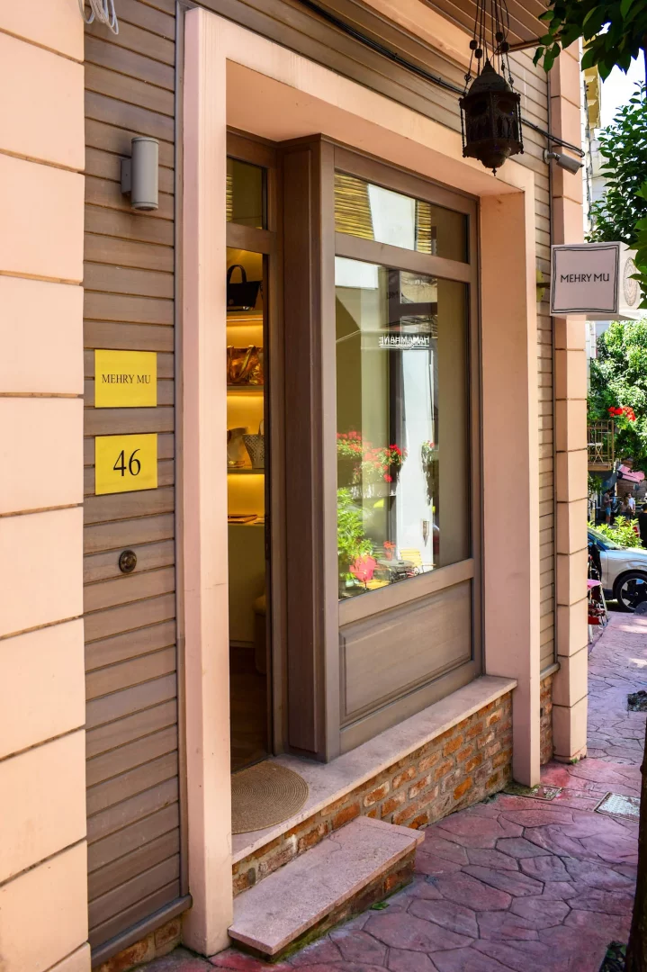 Çukurcuma'daki House of Mehry Mu butiğinin pastel pembe ve ahşap detaylı ön cephesi, kapısının solunda Mehry Mu ve 46 numaralı tabelaları, aydınlatılmış iç mekanda kişiselleştirilebilir çantaları sergileyen geniş vitrini ve zarif feneriyle zamansız tasarım anlayışını yansıtıyor.