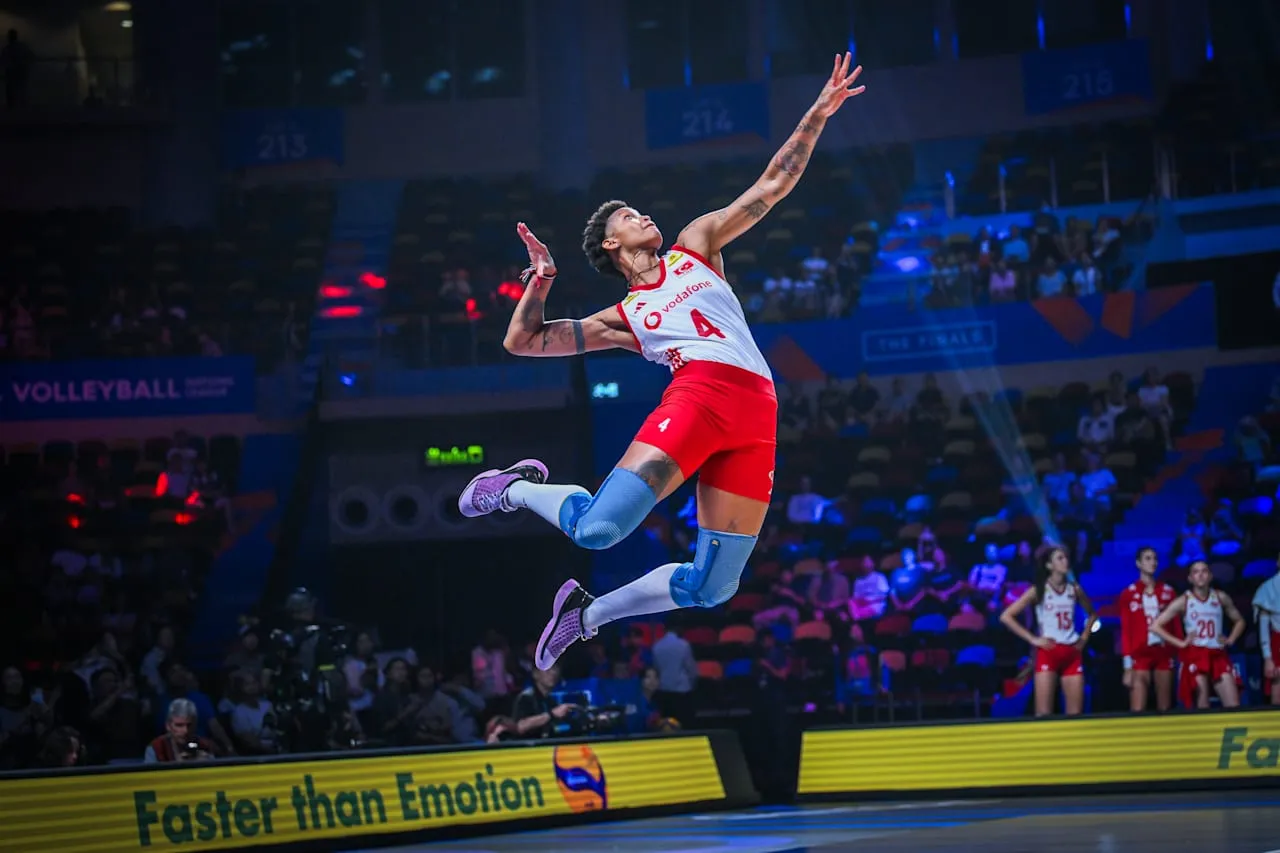 Melissa Vargas, 4 numaralı beyaz ve kırmızı forması, mavi dizlikleri ve mor spor ayakkabılarıyla bir voleybol maçında smaç vurmak üzere havada süzülürken görülüyor; kolları dövmelerle kaplı, odaklanmış bir ifadeyle topa bakıyor ve arkasında seyirci dolu bir spor salonu yer alıyor.