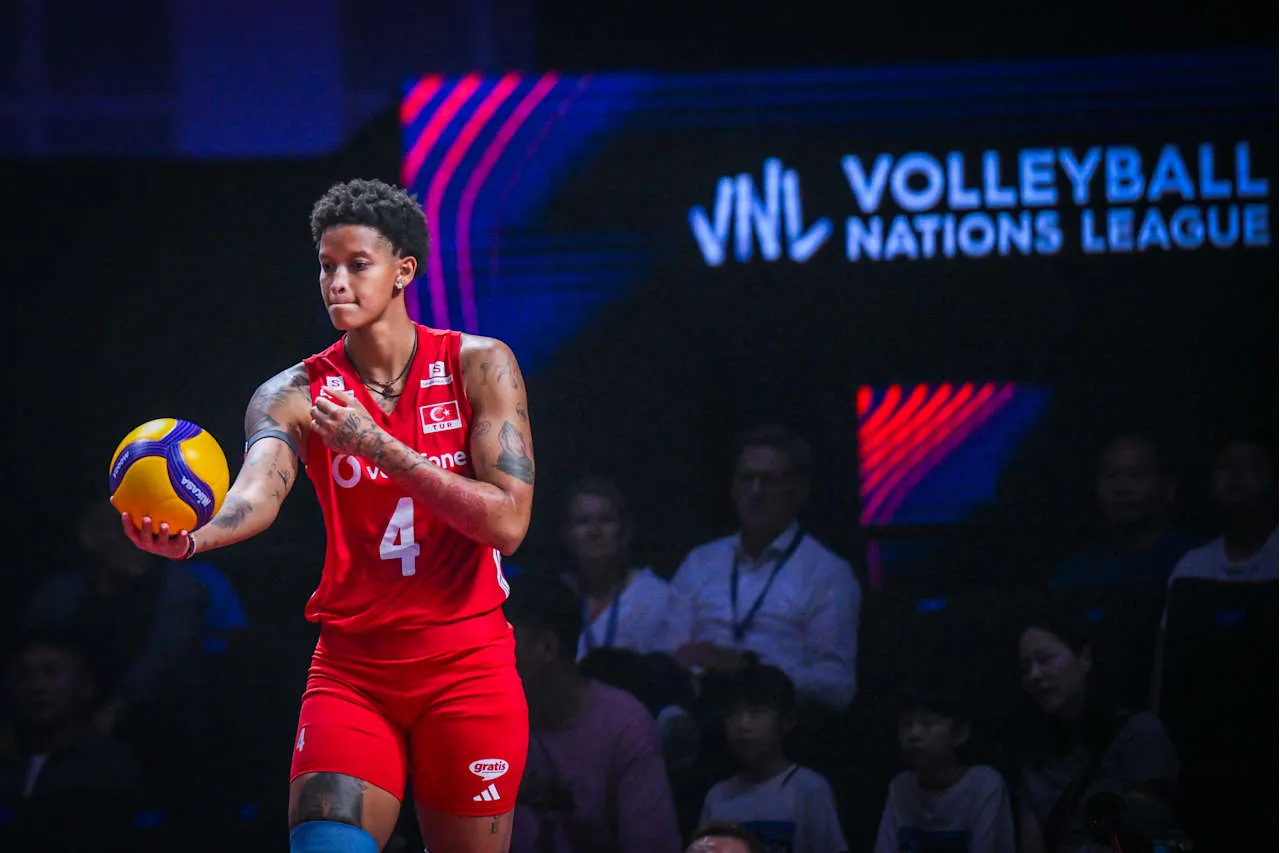 Voleybol oyuncusu Melissa Vargas, kırmızı milli takım forması ve kolundaki dövmeleriyle belirginleşerek, sarı-mavi bir voleybol topunu sol elinde tutarken, VNL Voleybol Milletler Ligi logosunun önünde ciddi bir ifadeyle sahada duruyor.