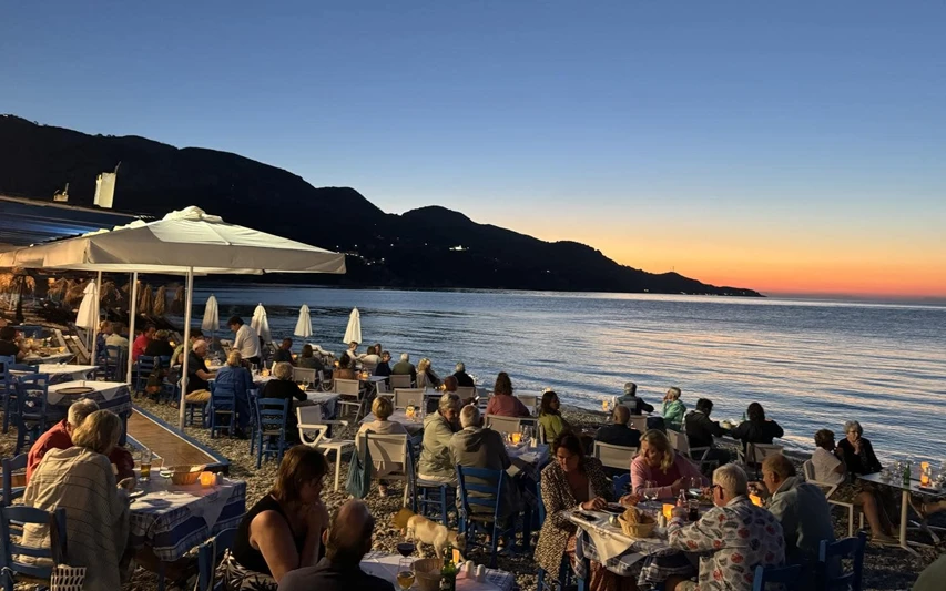 Samos'taki Meltemi Taverna'da, gün batımının ardından turuncu-mavi gökyüzü ve karanlık dağlar eşliğinde, denize sıfır çakıllı plajdaki mavi beyaz masa örtülü masalarda yemek yiyen ve sohbet eden kalabalık bir misafir grubu keyifli bir akşam geçiriyor.