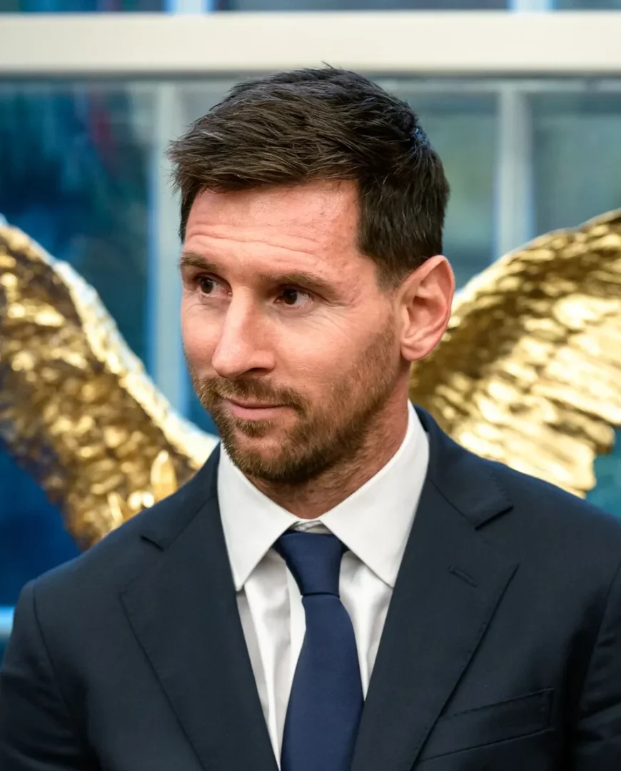 Koyu takım elbisesi, beyaz gömleği ve lacivert kravatıyla resmi bir duruş sergileyen Lionel Messi, hafifçe sola bakarken, uzun ömürlülüğünü ve başarılarını temsil eden arka plandaki parlak altın rengi kanat benzeri bir yapının önünde görülüyor.