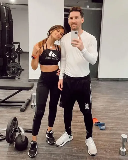 Lionel Messi'nin eşi Antonella Roccuzzo ile spor salonunda çekilen bir özçekimde, Messi beyaz uzun kollu bir spor tişörtü ve siyah şort giyerken, Antonella siyah spor sütyeni ve taytıyla gülümseyerek eşinin omzuna yaslanıyor ve bu kare, çiftin sağlıklı yaşam tarzı ve Messi'nin spor kariyerindeki uzun ömürlülüğü için fiziksel dayanıklılığın önemini vurguluyor.