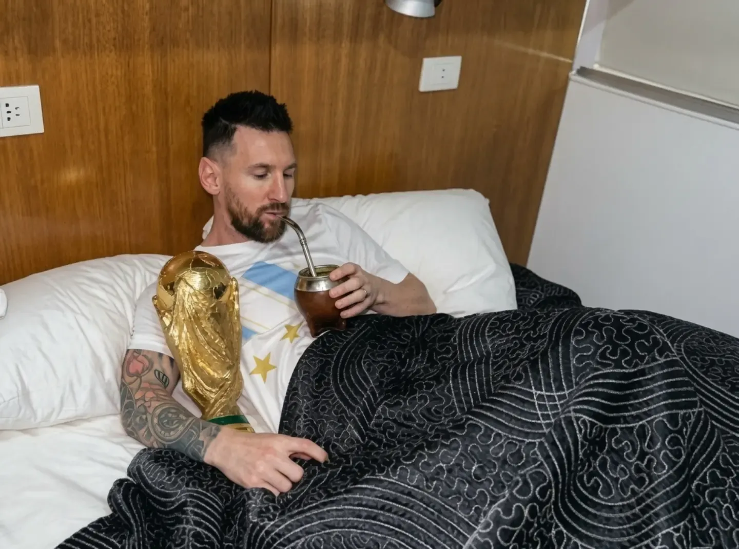 FIFA Dünya Kupası'nı kazanmanın ardından yatağında uzanırken, kupa kollarının arasında ve elinde bir mate içeceği ile keyifli bir an yaşayan Lionel Messi'nin, profesyonel spor hayatındaki uzun ömürlülük ve beslenme düzeninin başarısındaki rolünü yansıtan bir görsel.