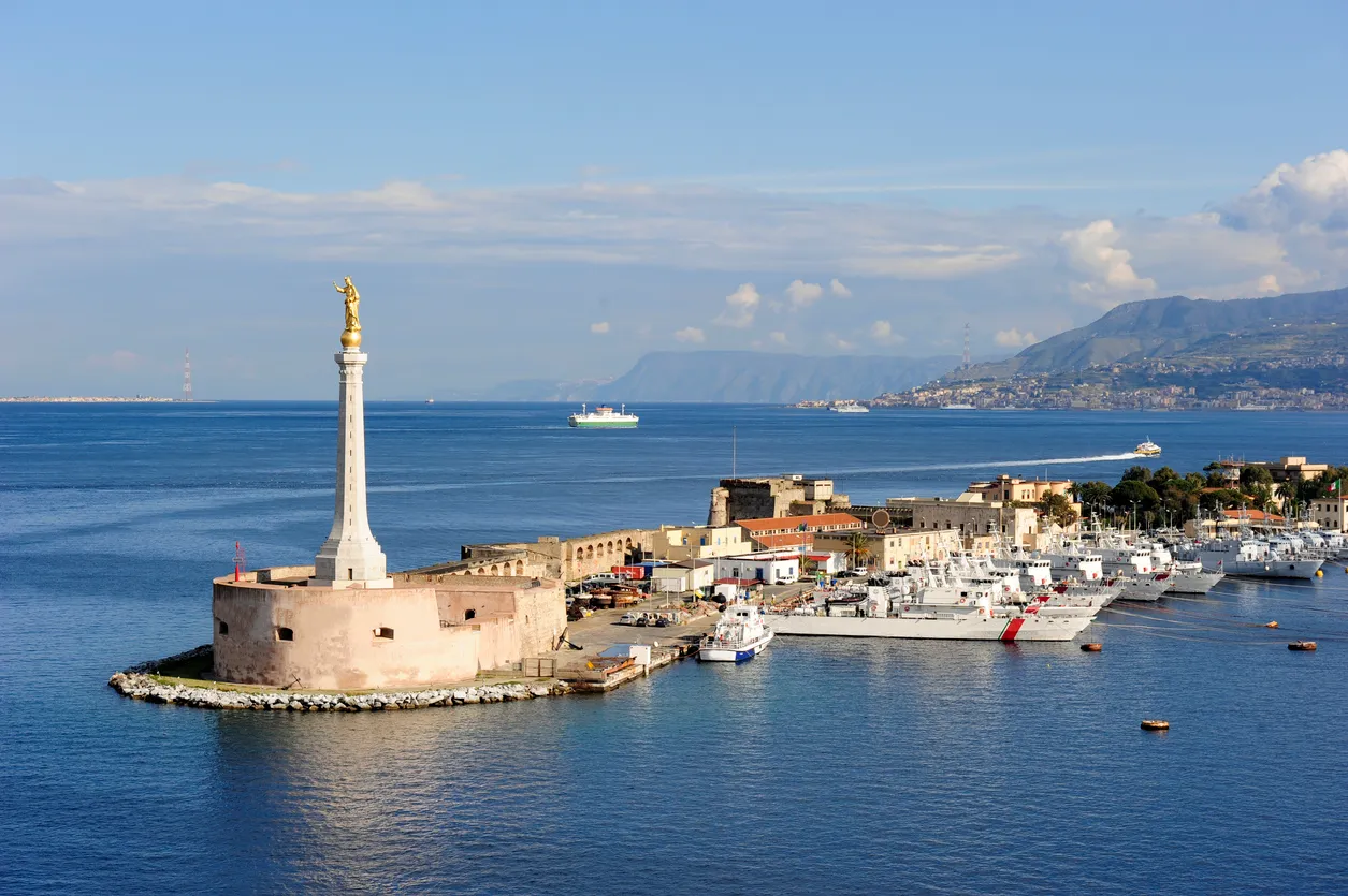 Messina Boğazı'nın mavi suları üzerinde, altın rengi bir heykelle taçlandırılmış yüksek beyaz sütunun ve etrafındaki tarihi yapının yer aldığı burun, sağ tarafta sıralanmış çok sayıda beyaz gemi ve binalarla çevrili hareketli bir liman alanını gösteriyor; ufukta dağlar, uzakta görünen şehir ve ilerleyen gemilerle boğazın genel görünümü vurgulanıyor.