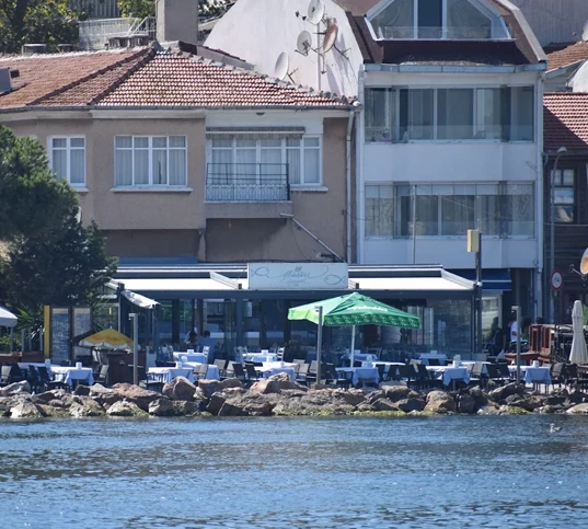 Kınalıada'daki Mimoza Restoran, deniz kenarında, masaları beyaz örtülerle donatılmış ve bazıları yeşil şemsiyelerle gölgelendirilmiş açık hava oturma alanıyla dikkat çekiyor; önünde kayalık bir kıyı şeridi bulunan bu huzurlu mekan, sakin sulara nazır keyifli bir yemek deneyimi sunuyor.