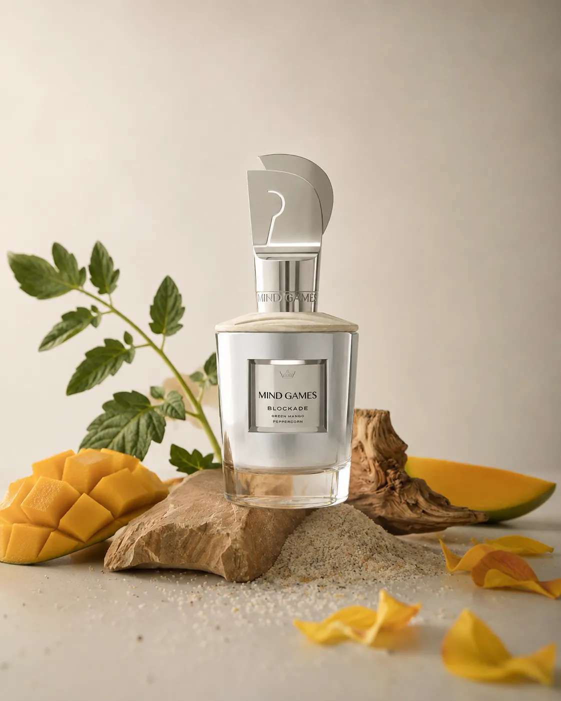 Mind Games Blockade erkek parfümü şişesi, üzerinde Green Mango Peppercorn yazan etiketiyle, keskin hatlı gümüş renkli soru işareti şeklindeki kapak detayıyla, küp kesilmiş mango dilimleri, yeşil yapraklı bir dal, kumlu zemin ve kuru ağaç parçacıkları arasında zarif bir şekilde konumlandırılmış.