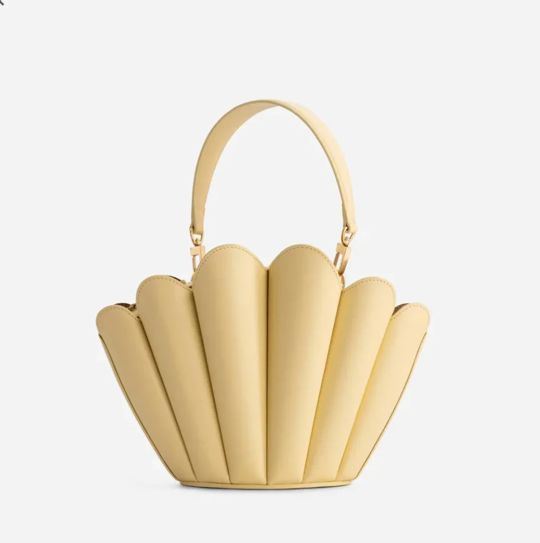 Açık tereyağı rengindeki bu MlOUYE deniz kabuğu formunda tasarlanmış, altın rengi metal aksesuarlara sahip ve hem elde hem de çapraz askılı (crossbody) olarak kullanılabilen zarif çanta, ürünün detaylarını net bir şekilde gösteren beyaz bir arka planda sergileniyor.
