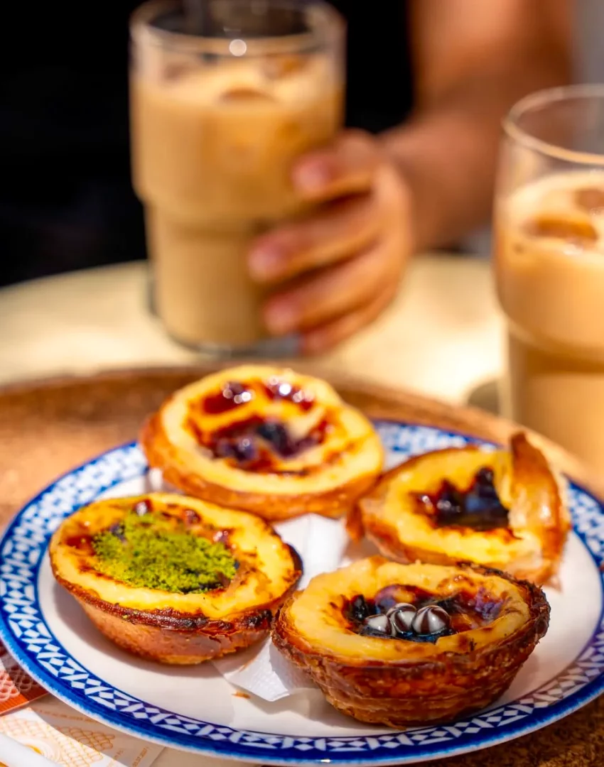 Mavi desenli beyaz bir tabakta, üzeri fıstık, yaban mersini ve çikolata parçacıklarıyla süslenmiş dört adet farklı çeşit Pastel de Nata Portekiz tatlısı, arka planda bir elin tuttuğu buzlu kahve ile birlikte güneşli bir kafe masasında yer alıyor.