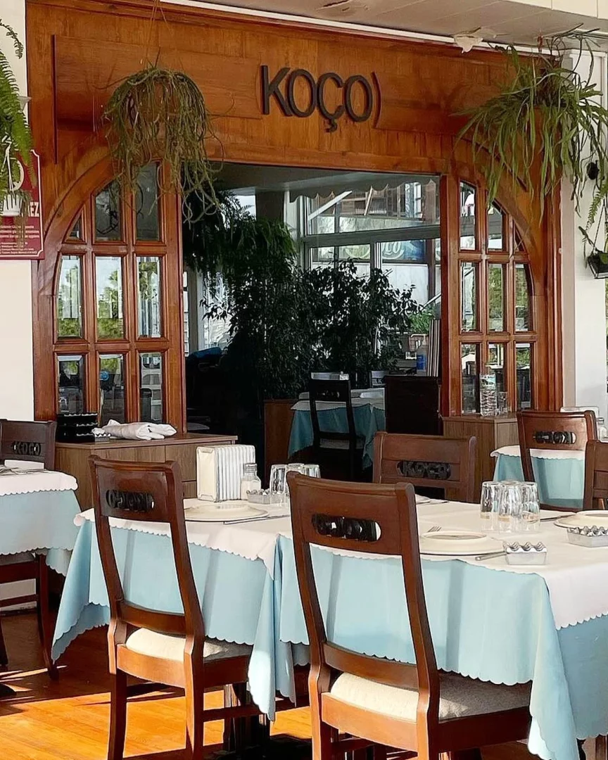 Moda'nın tarihi ve klasik lezzet duraklarından Koço Restoran'ın iç mekanında, ahşap detaylar, geniş pencerelerle aydınlanan ferah bir yemek alanı, asılı yeşil bitkiler, beyaz ve açık mavi masa örtüleriyle hazırlanmış boş masalar ile koyu renk ahşap sandalyeler görülüyor.