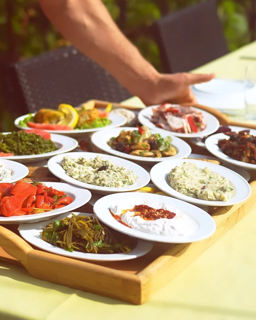 Moda Koço Restoran'ın güneşli bir açık hava ortamında, sarı masa örtüsü serili bir masaya, bir garsonun eliyle servis edilen, ahşap tepside özenle sunulmuş birbirinden çeşitli ve taze Türk mezeleri iştah açıcı görünüyor.