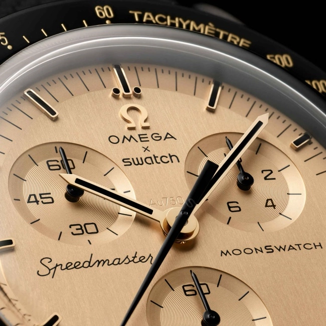 Ay Görevi temalı sınırlı üretim Moonswatch kol saatinin altın saten kadran detayında, parlak siyah çerçevesi üzerinde TACHYMÈTRE yazısıyla, ortada OMEGA X SWATCH logosu, altında Au750, Speedmaster ve MOONSWATCH yazıları ile siyah ibreler ve üç kronograf alt kadranı belirgin şekilde görülüyor.