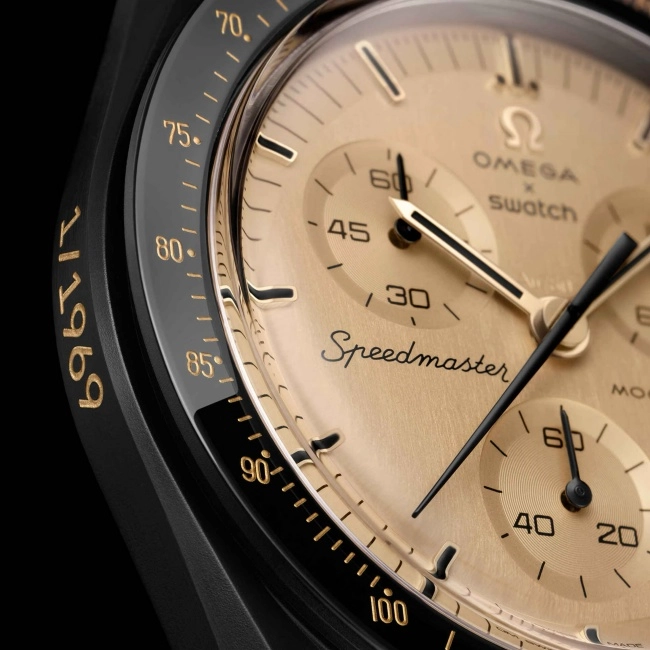 Siyah kasalı ve detaylı altın rengi fırçalanmış kadrana sahip, Moonswatch Mission to the Moon sınırlı üretim kol saatinin bu yakın çekim görüntüsünde, kadran üzerinde belirgin Speedmaster yazısı, Omega x Swatch logoları ve yan bezelde altın rengi 1969 yılı ile takimetre ölçeği açıkça görünmektedir.